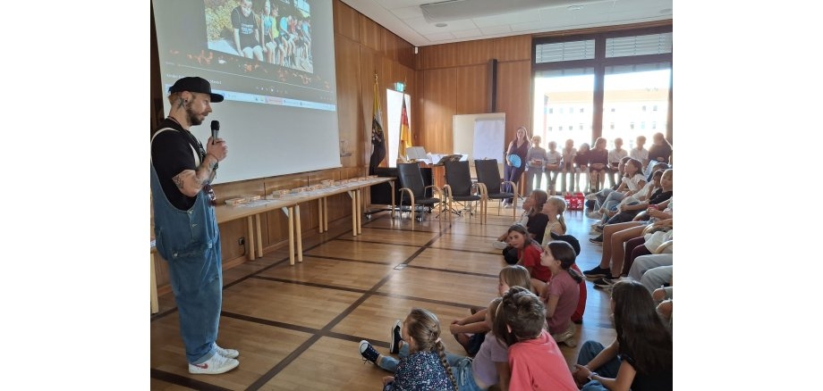Eine Gruppe von Kindern sitzt im Halbkreis auf dem Boden vor einer Leinwand im Sitzungssaal der Kreisverwaltung. Ein Mann mit Baseballkappe hat ein Mikrofon in der Hand und moderiert die Veranstaltung.