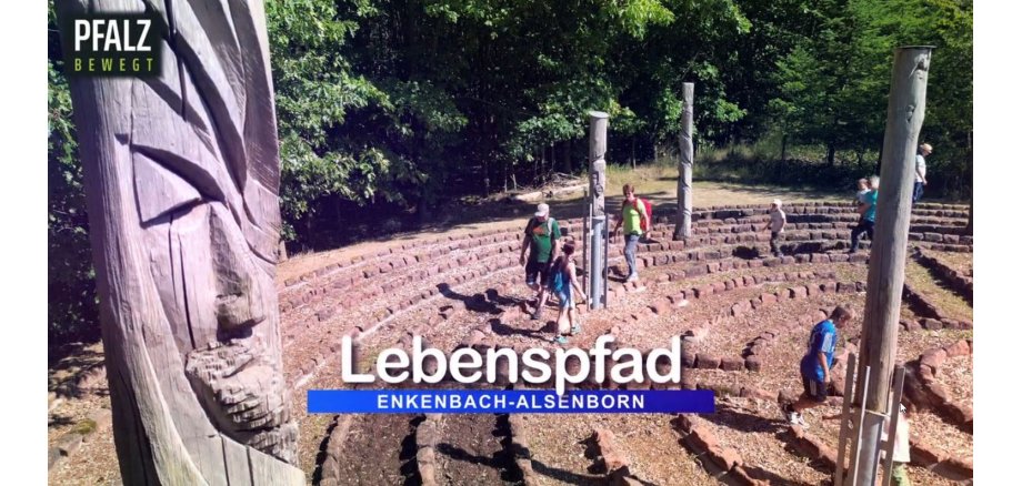 Man sieht einen Ausschnitt des Labyrinths am Lebenspfad. Am linken Bildrand sieht man eine Skulptur aus einem Holzstamm geschnitzt. Im Hintergrund laufen mehrere Menschen (Frauen, Männer, Kinder) durch die Gänge des Labyrinths.